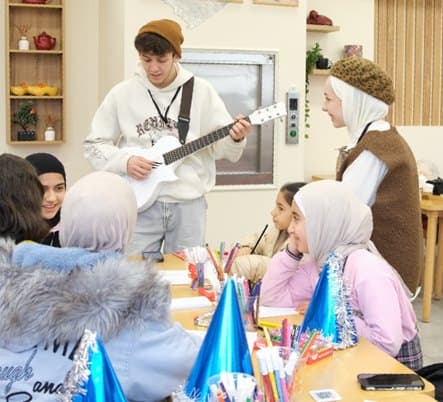 Gitar çalan bir gönüllü ve parti şapkalı çocuklarla dolu atölye masası