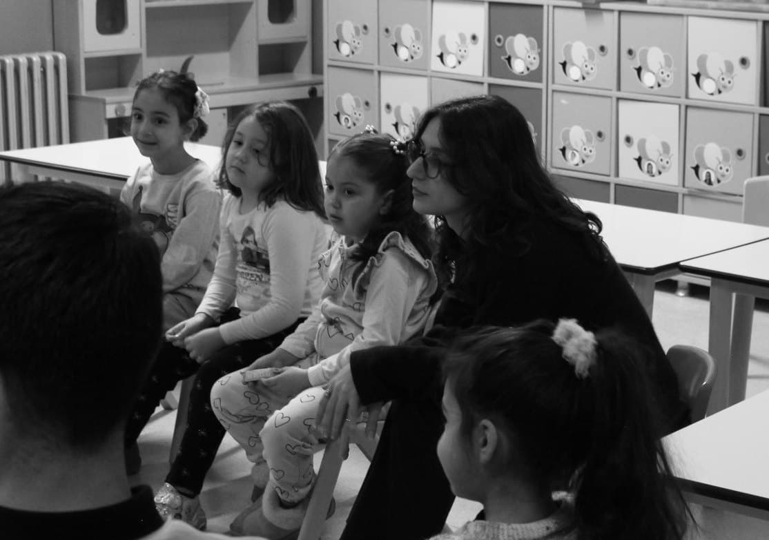 Bir sınıfta yan yana oturmuş küçük çocuklar ve onları dinleyen bir Sketch gönüllüsü
